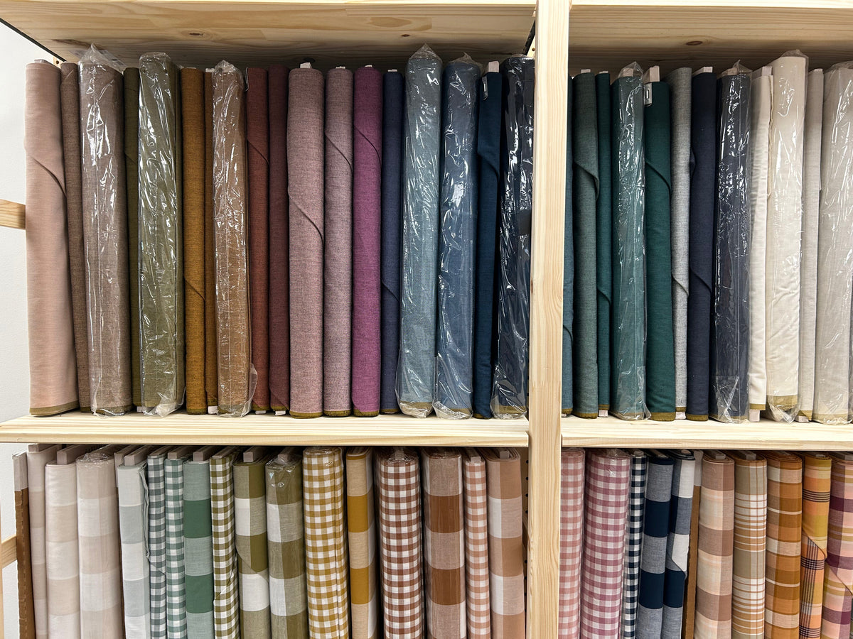 Wooden shelves stocked with rolled fabrics in many colors and patterns, including solid tones, blues, greens, plaid, and gingham, many wrapped in clear protective plastic.