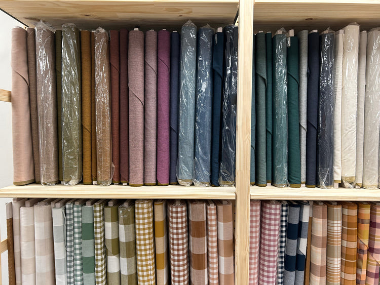 Wooden shelves stocked with rolled fabrics in many colors and patterns, including solid tones, blues, greens, plaid, and gingham, many wrapped in clear protective plastic.