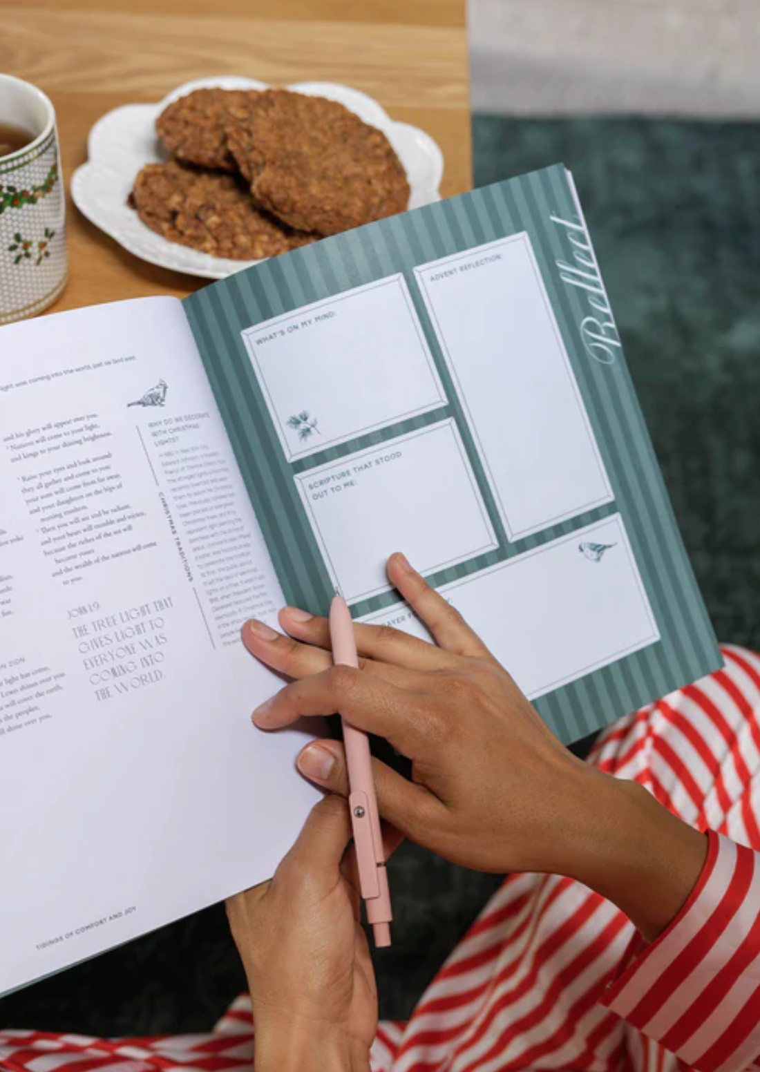 Person holding a notebook with a pen, next to a plate of cookies and a cup on a wooden table.